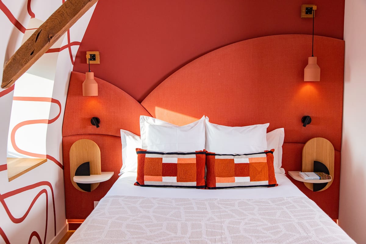 Une chambre d’hôtel moderne avec une tête de lit rouge incurvée, du linge de lit blanc et deux tables de chevet sur lesquelles sont posés des livres.