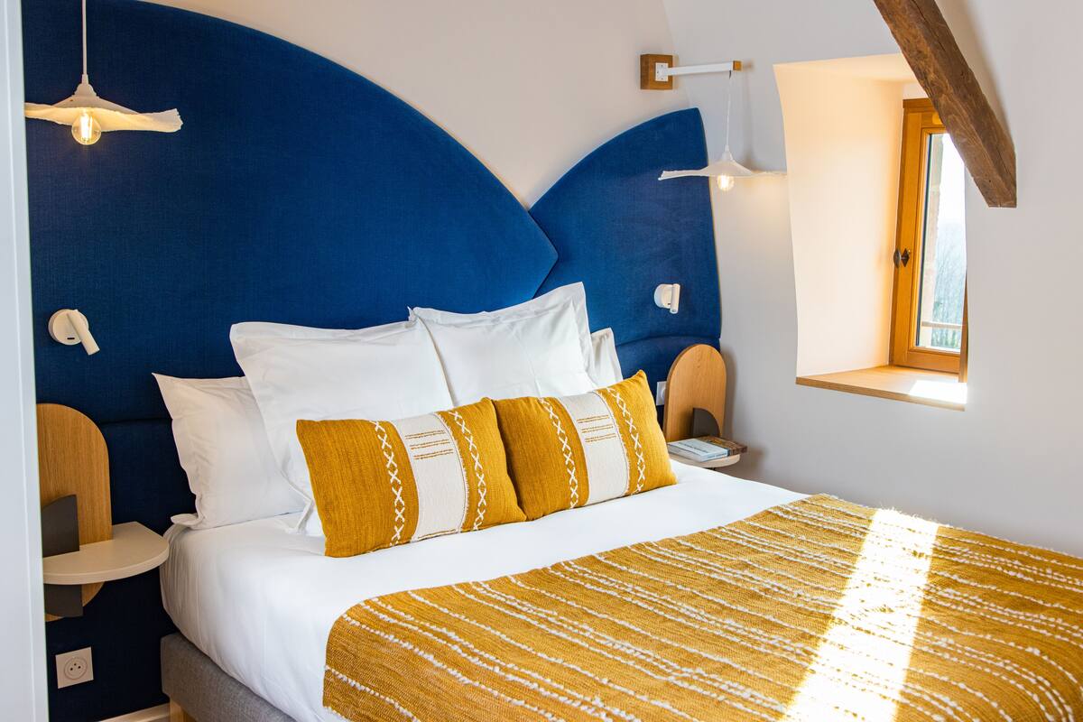 Une chambre avec un grand lit, une tête de lit bleue, des coussins blancs et moutarde, une couverture jaune à rayures, des tables de chevet et une fenêtre avec vue.