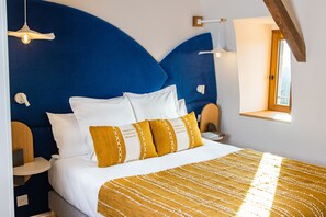 A bedroom with a large bed, blue headboard, white and mustard pillows, a yellow striped blanket, bedside tables, and a window with a view. - Cueillette (Altillac)