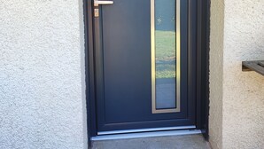 Eine dunkel gefärbte Tür mit einem vertikalen Glasschieber, einer Metallglocke und einem kleinen Holzregal zur Rechten.