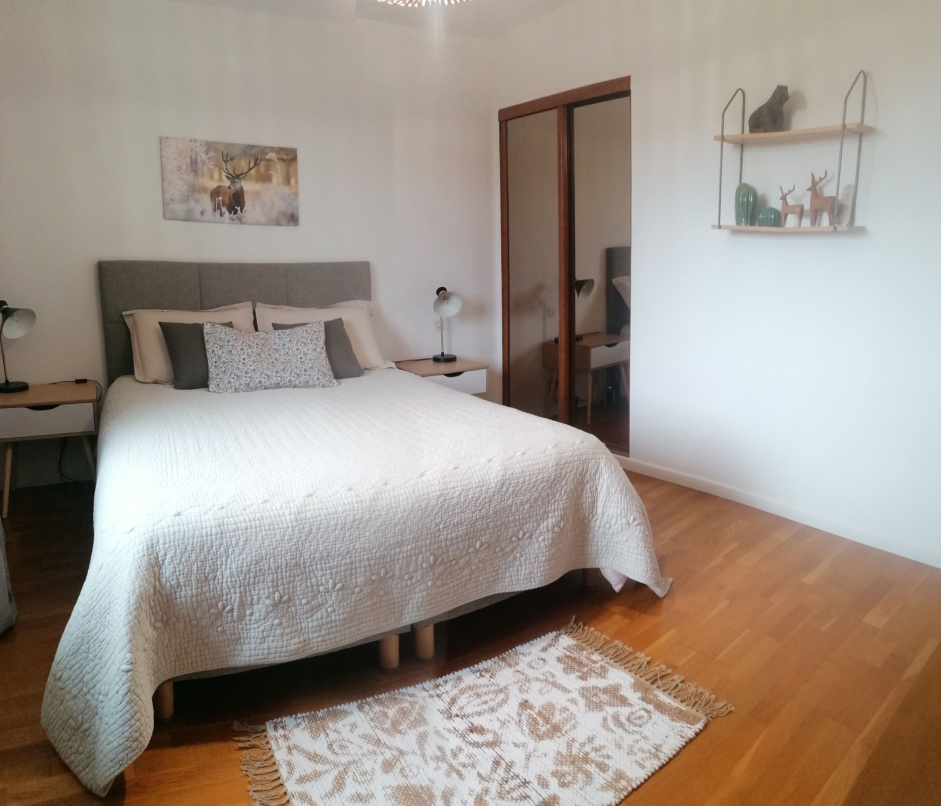 A bedroom with a large bed, a bedside table with a lamp, a nightstand, a wooden floor, a patterned rug, a sliding door, and wall decorations including a deer and a shelf with items.