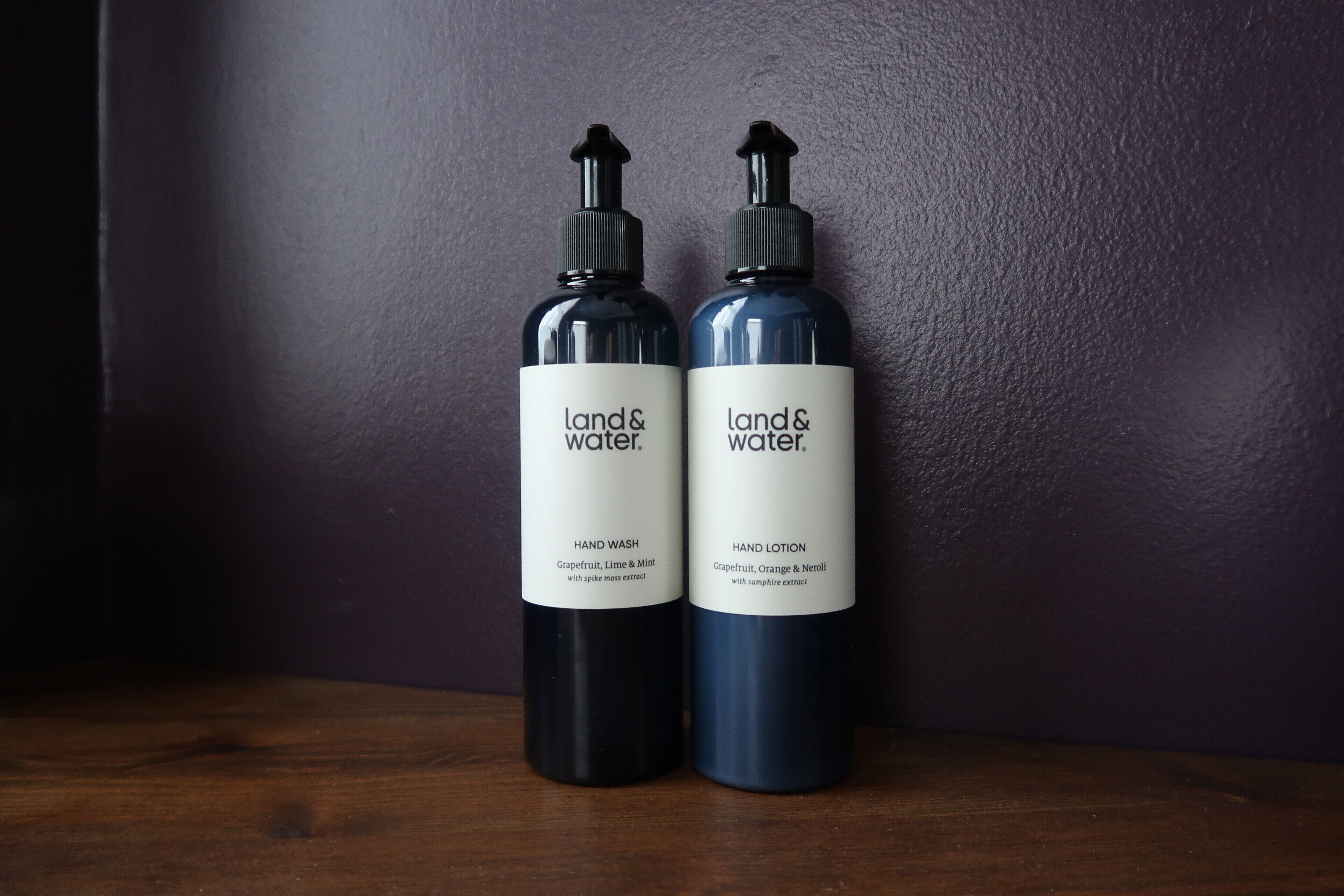 Two bottles of 'Land & Water' hand wash and hand lotion on a wooden surface against a purple wall.