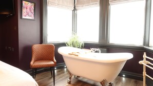 A bathroom with a freestanding bathtub, a cushioned chair, a potted plant, and a large window with blinds. - The Craft House (Plymouth)