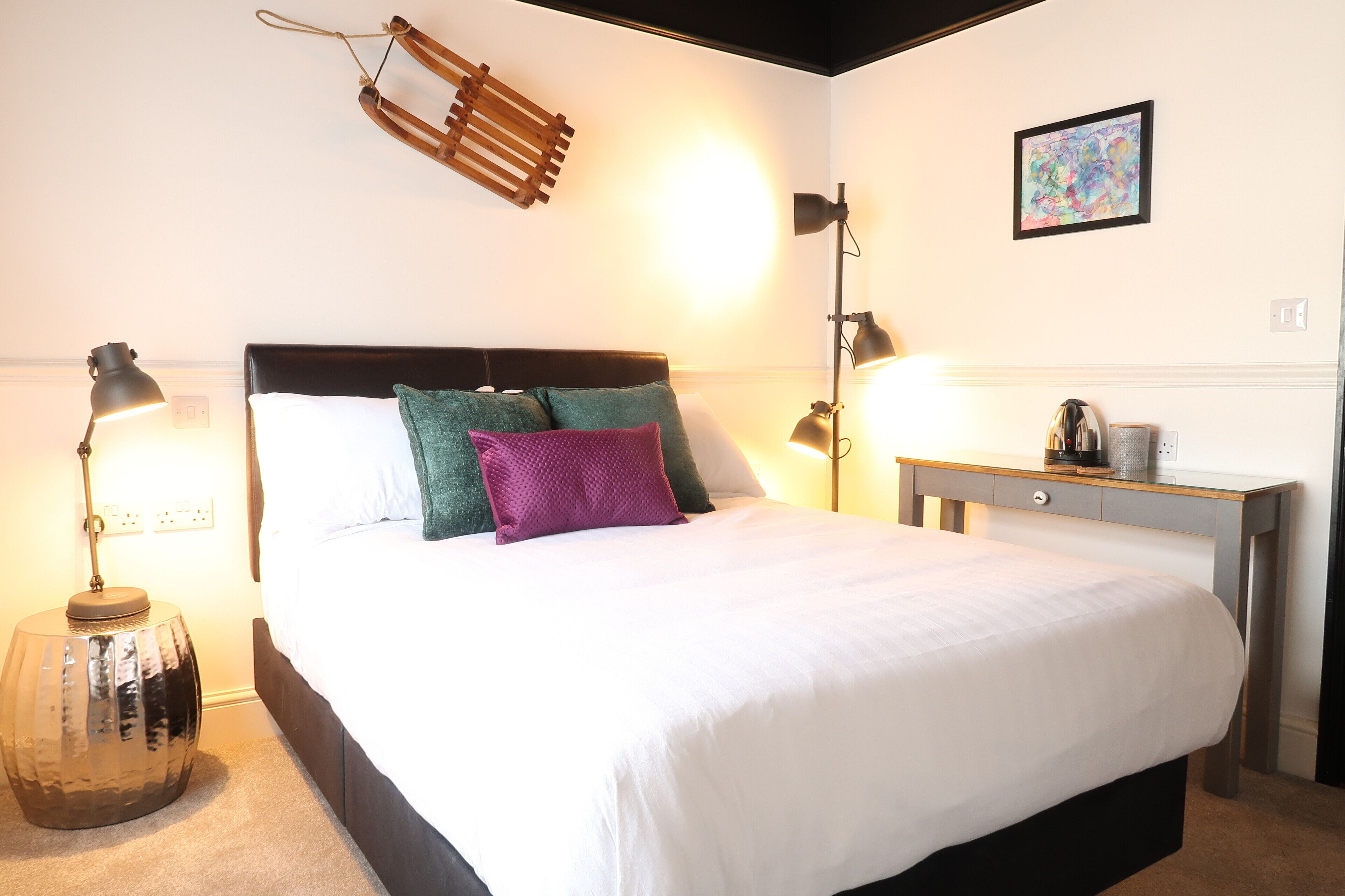 Een net opgemaakt bed met witte beddengoed, een hoofdbord en twee kussens. Er staat een nachtkastje met een lamp en een klein decoratief object.