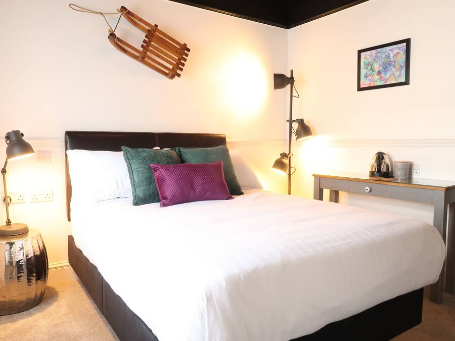 Een net opgemaakt bed met witte beddengoed, een hoofdbord en twee kussens. Er staat een nachtkastje met een lamp en een klein decoratief object.