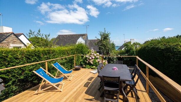 A wooden deck with a table and chairs, surrounded by greenery and a view of houses.