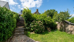 A stone pathway leading to a traditional stone house with a wooden gate, surrounded by lush greenery and flowering bushes.