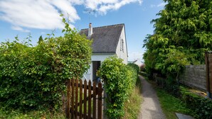 A narrow gravel path leading to a white house with a dark roof, surrounded by lush greenery and a wooden fence.