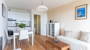 Una sala moderna con un sofá blanco, una mesa de centro de madera y un comedor con una mesa y sillas blancas. Se ve una cocina con armarios blancos y un colgante en el fondo.