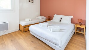 Un dormitorio con una cama grande, una cama más pequeña, dos mesitas de noche con lámparas, un cuadro en la pared y una ventana.