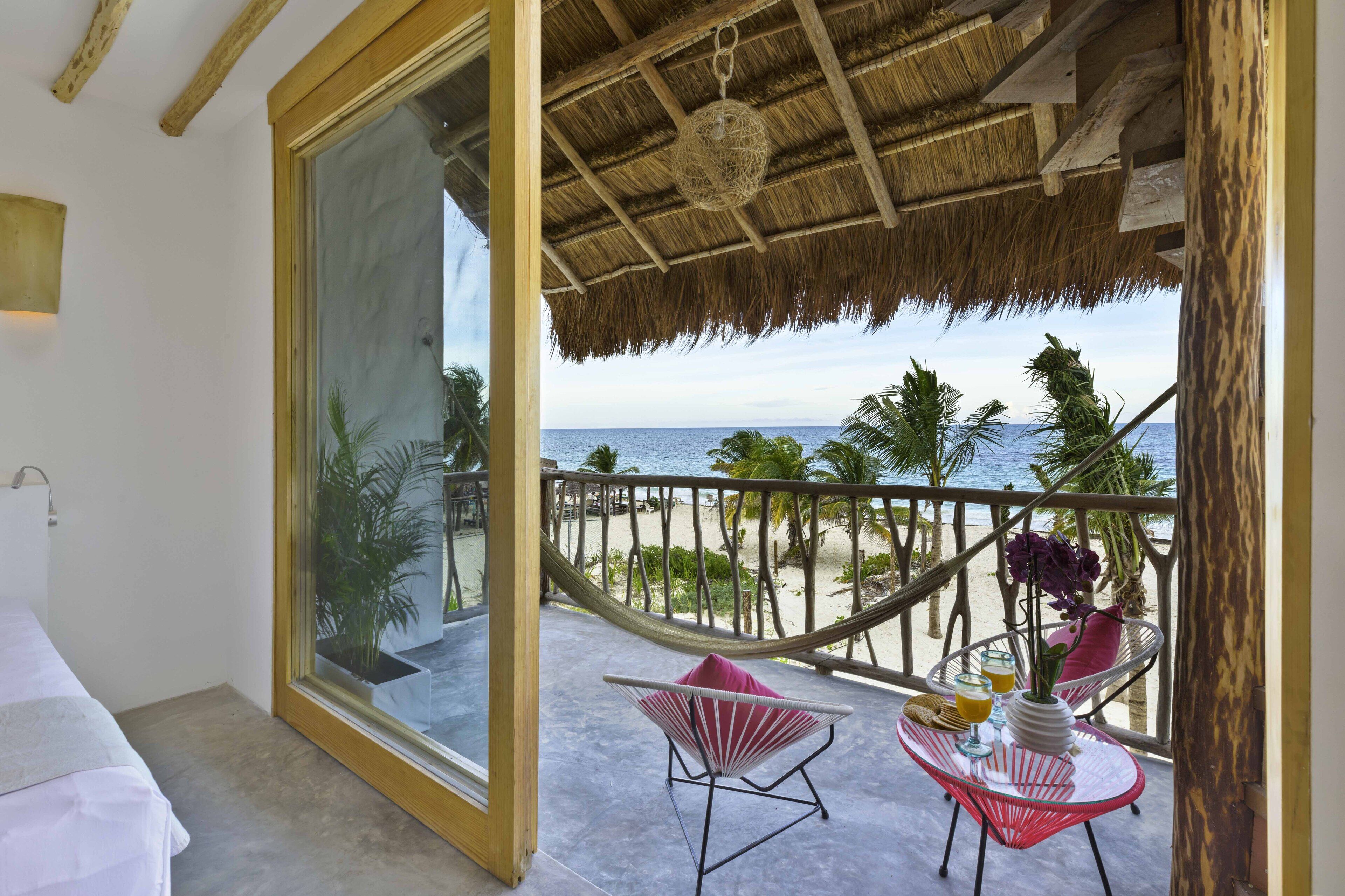 A balcony with a view of the beach, a hammock, a chair, and a table with drinks and a vase.