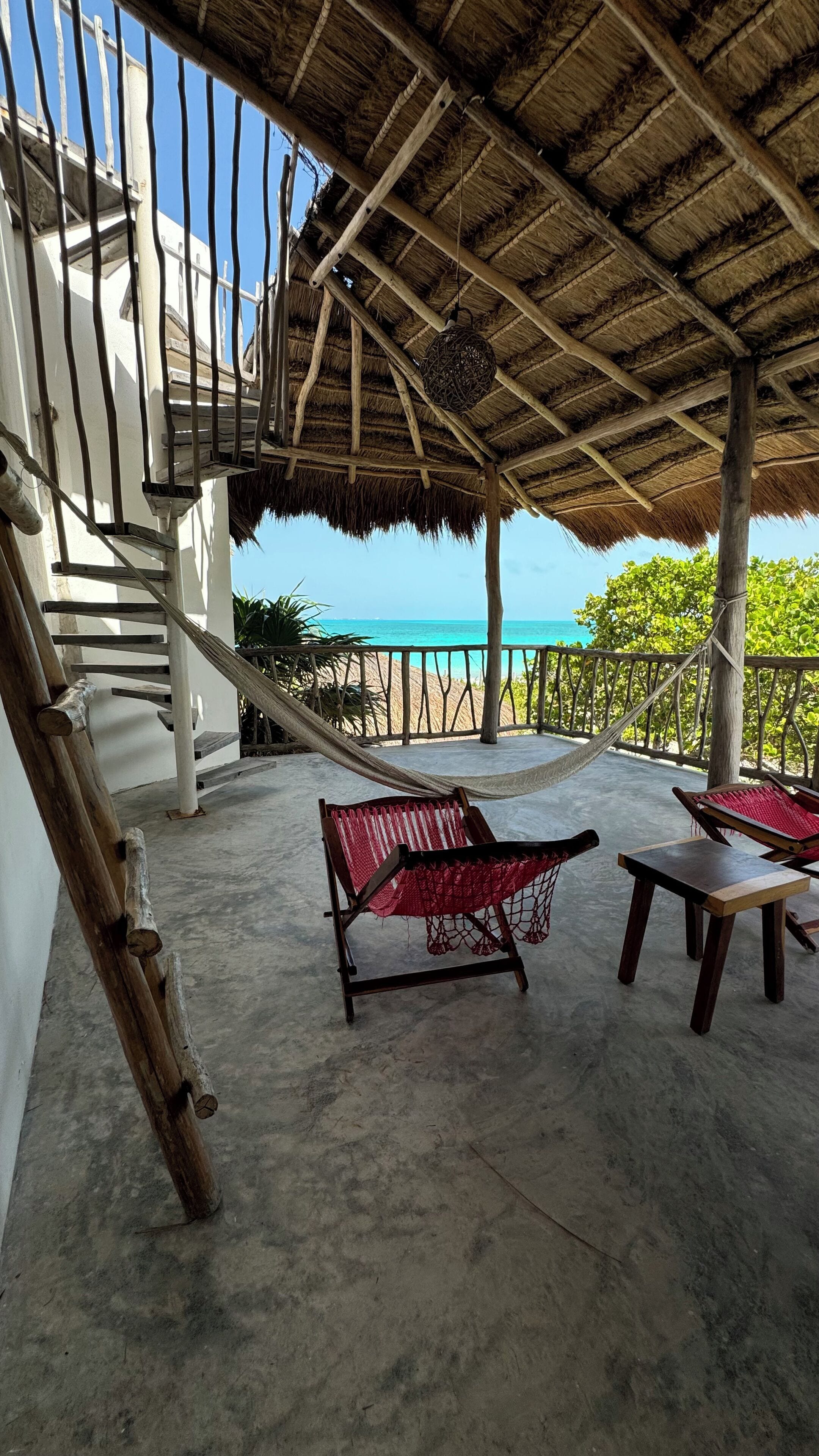 A covered outdoor area with a hammock, two wooden chairs, and a staircase leading to a balcony overlooking the sea.