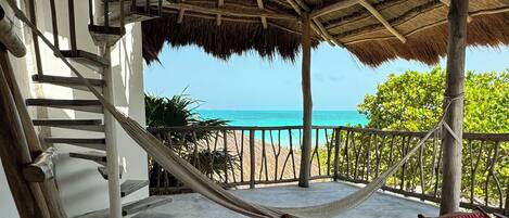 A covered outdoor area with a hammock, two wooden chairs, and a staircase leading to a balcony overlooking the sea.