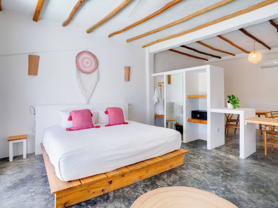 A modern bedroom with a wooden platform bed, a small bedside table, a wall-mounted decorative item, and a dining area with a table and chairs.