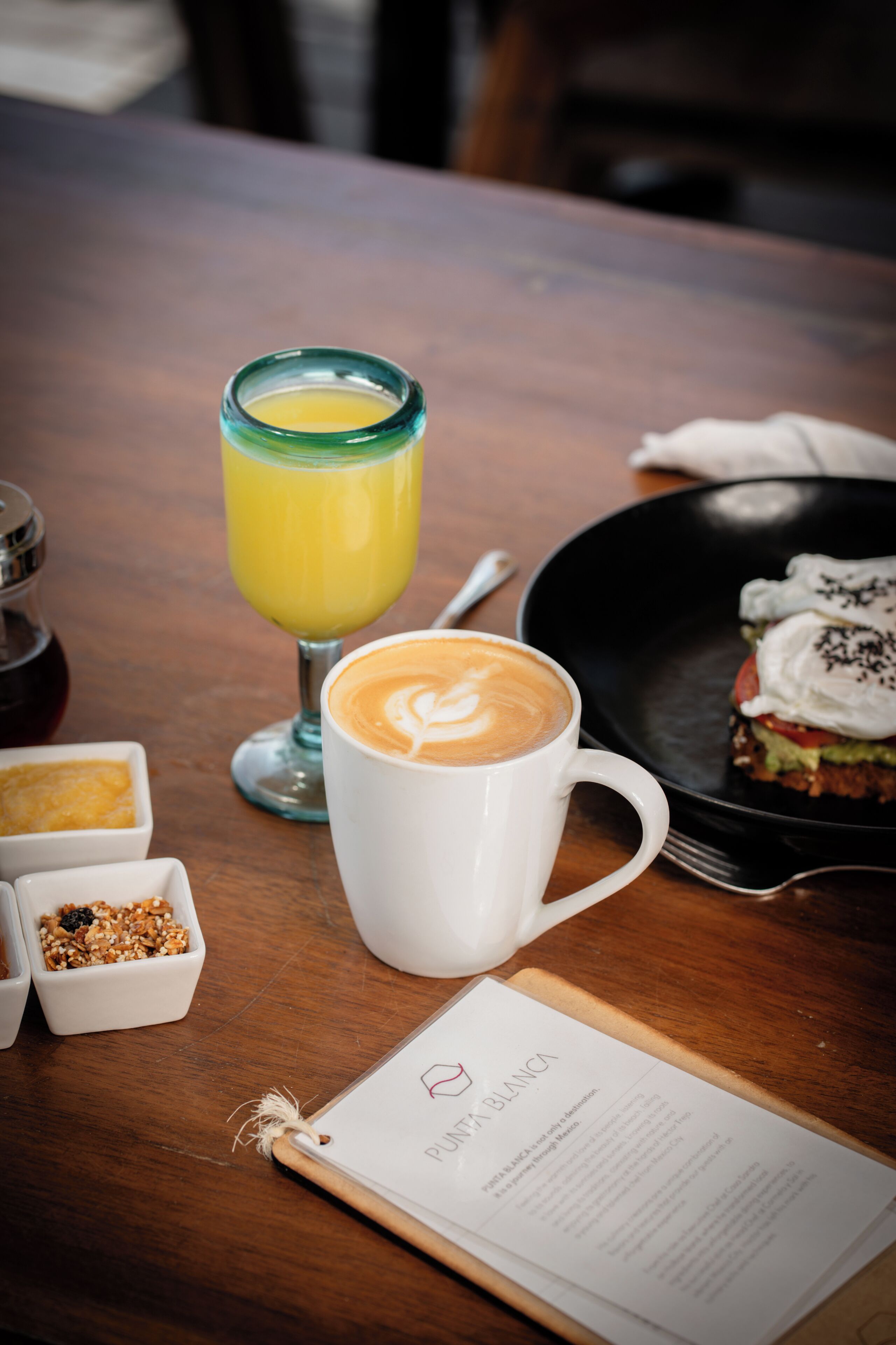 Ein Holztisch mit einer weißen Tasse, einem Glas Orangensaft, einem Teller mit einem Sandwich und einer Speisekarte.