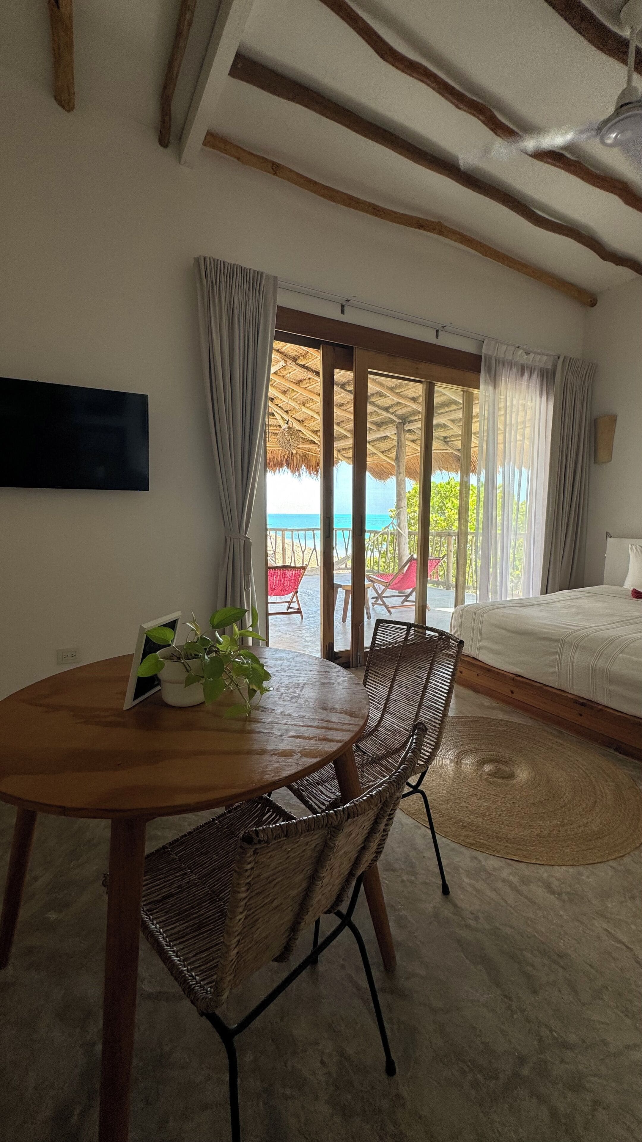 A room with a wooden table and wicker chairs, a bed with a white duvet, and a view of the beach through a glass door.