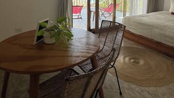 A room with a wooden table and wicker chairs, a bed with a white duvet, and a view of the beach through a glass door.