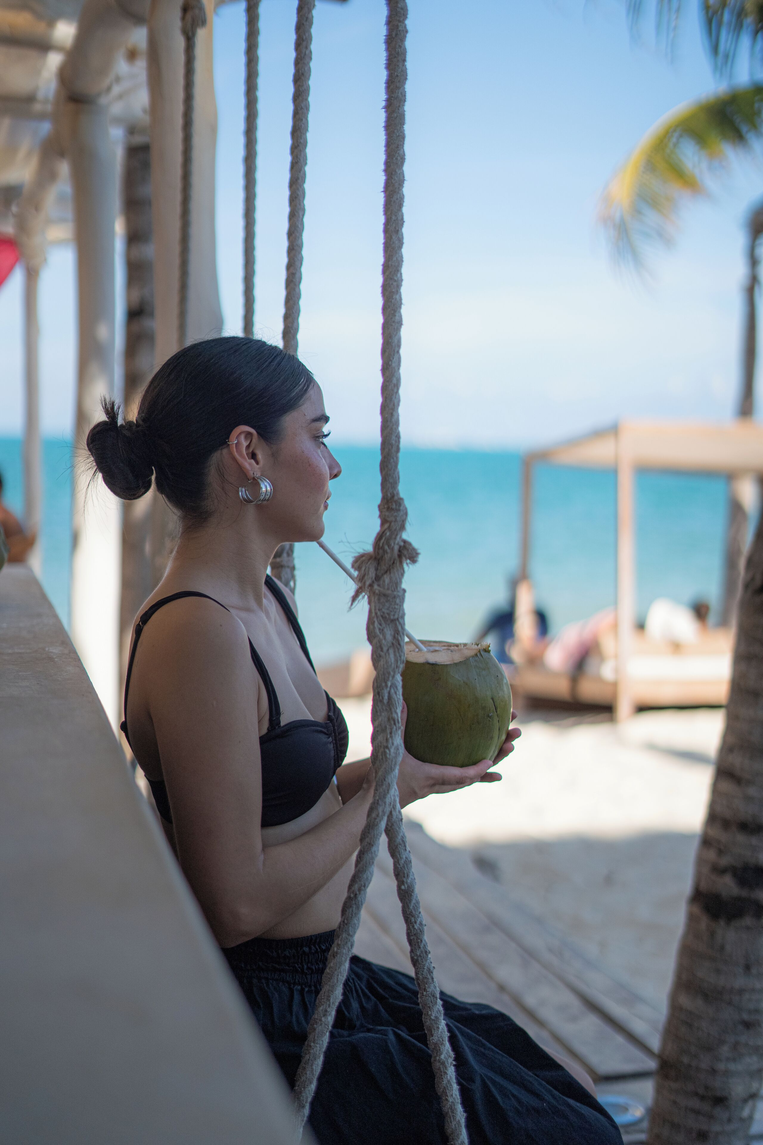 A person sitting on a wooden deck holding a coconut, with a beach and ocean in the background.