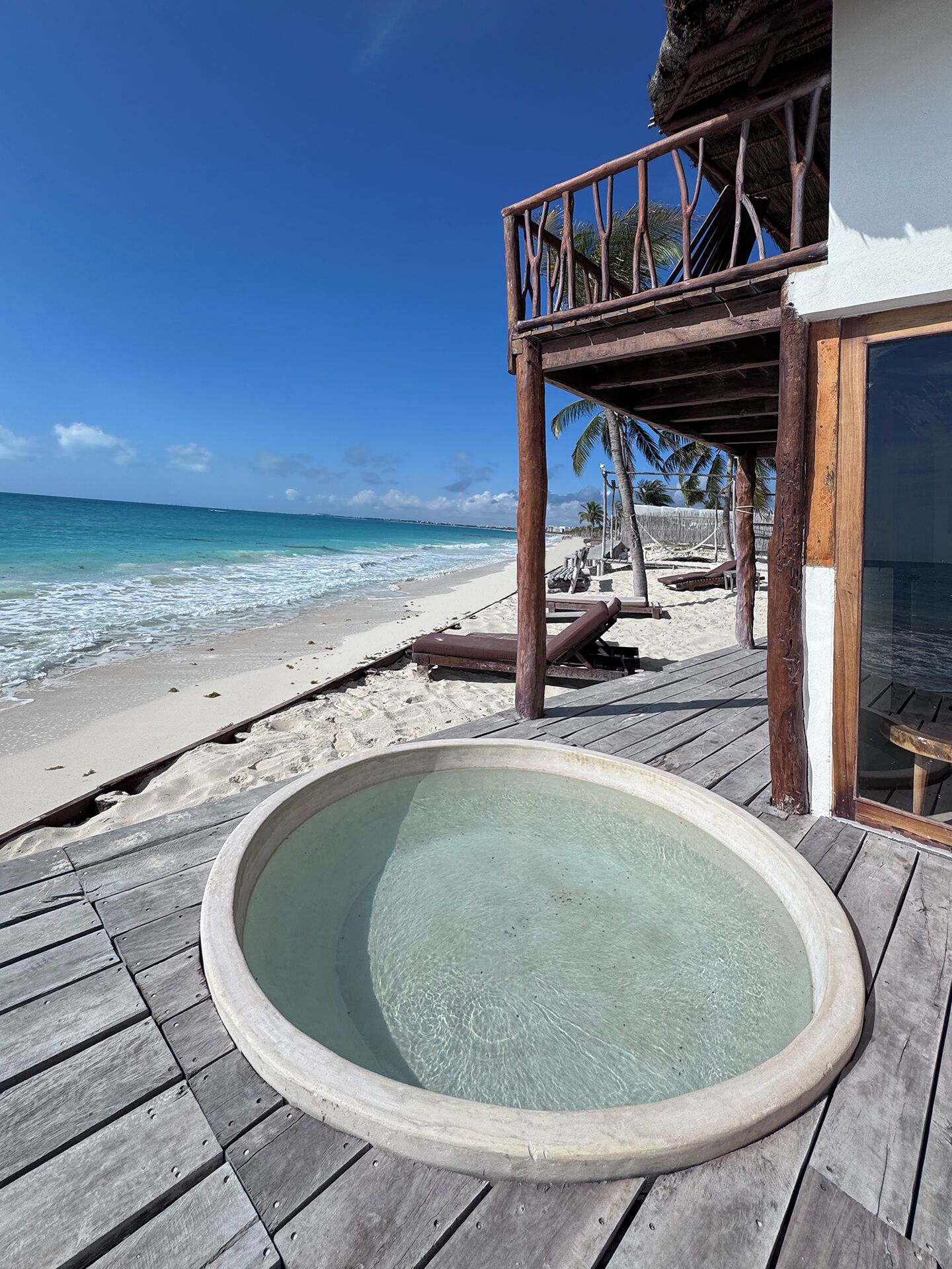 A wooden deck with a hot tub, overlooking a beach with palm trees and a clear blue sky.