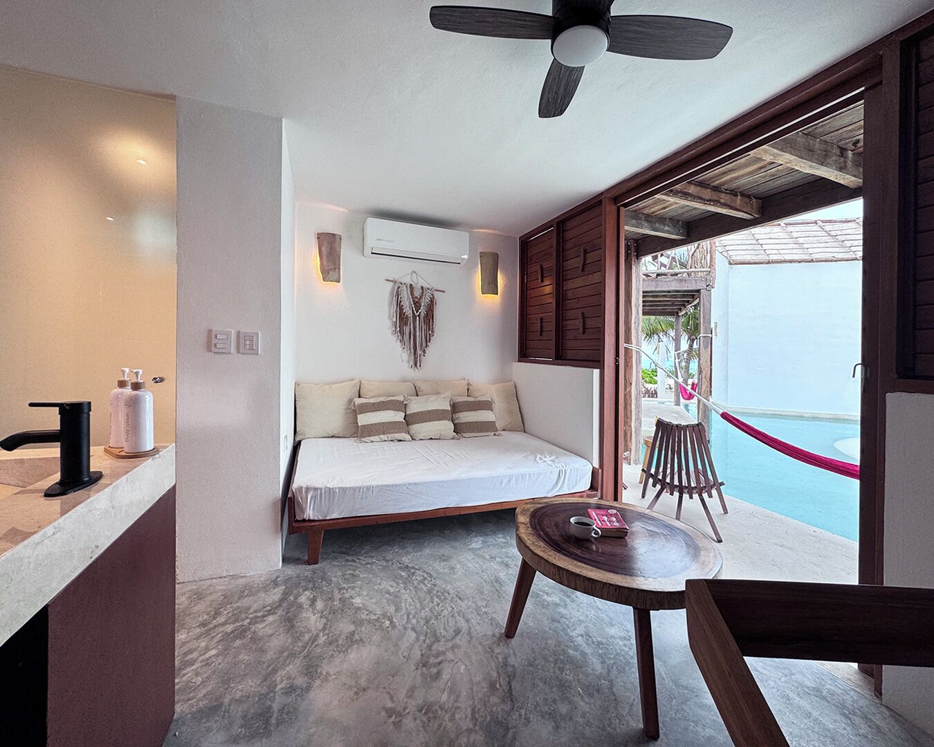 Un dormitorio con cama, ventilador de techo, una pequeña mesa redonda con una taza y un libro, y vista a una piscina exterior.