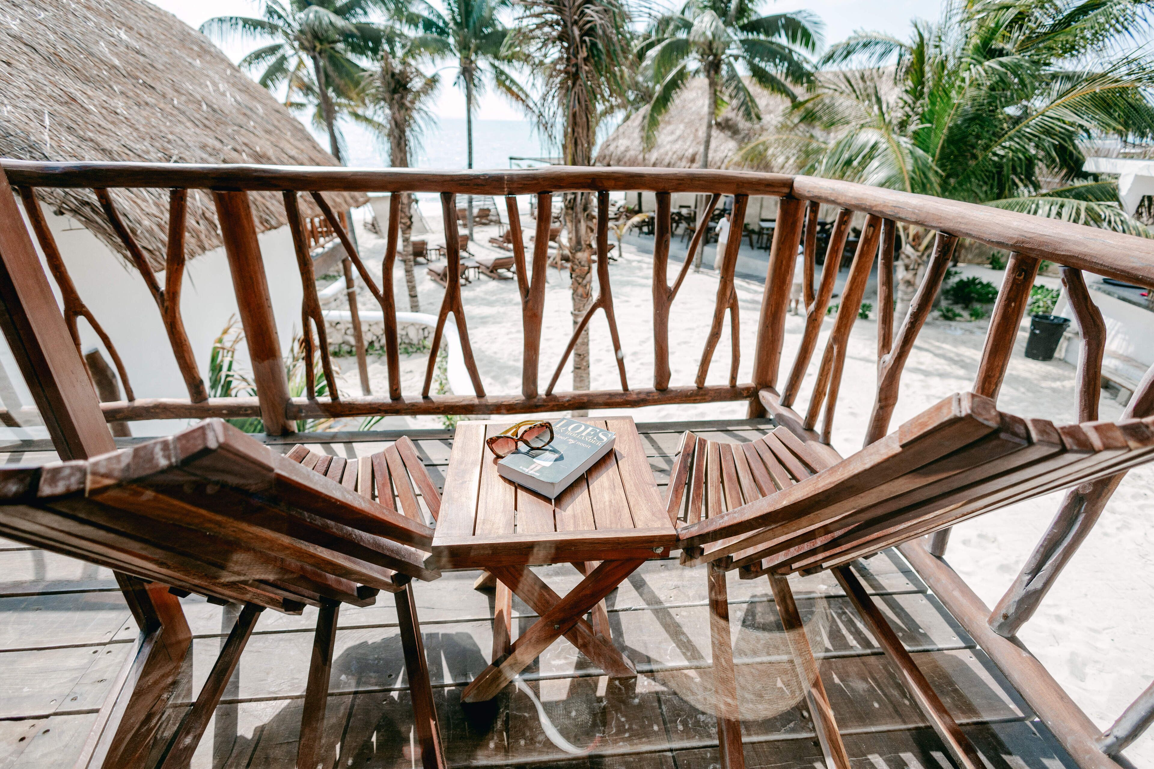 Un banco de madera con una mesita de vidrio, un libro y gafas de sol encima, situado en una terraza con vistas a palmeras y la playa.