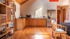 Une cuisine compacte avec des meubles en bois, un four à micro-ondes et un réfrigérateur. La pièce adjacente contient une chaise en bois et un lit recouvert d’une couverture foncée.
