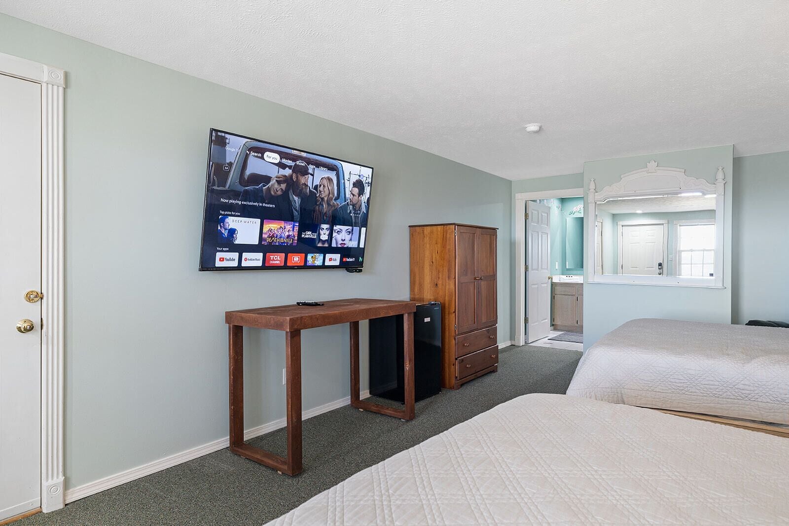 A hotel room with a television mounted on the wall displaying a movie, a wooden cabinet, a small table, and two beds.