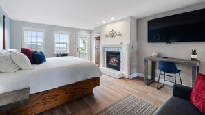 A modern bedroom with a large bed, a fireplace, a TV mounted on the wall, and a view of the outdoors through the windows. - The Bradford (Branson)
