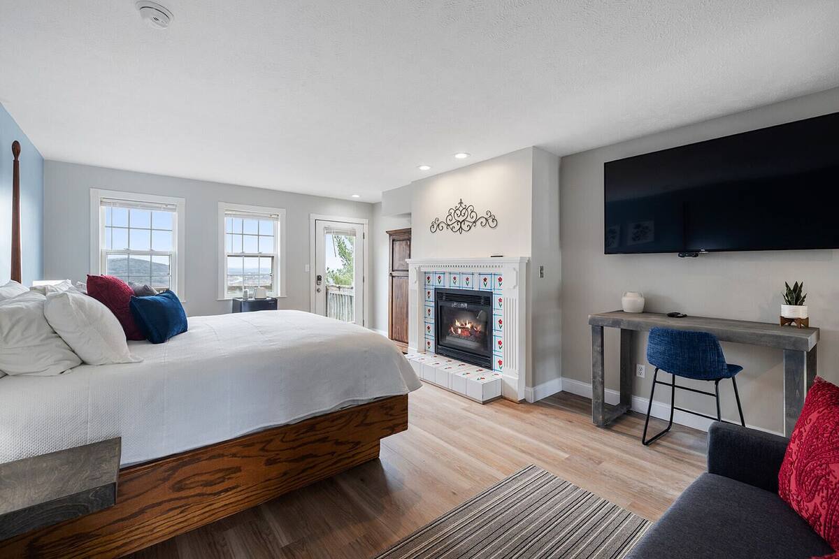 A modern bedroom with a large bed, a fireplace, a TV mounted on the wall, and a view of the outdoors through the windows.