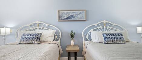 Ein Zimmer mit zwei Betten, jeweils mit weißem Metallgestell und gemusterten Kissen, einem kleinen Tisch mit einer Vase sowie einem gerahmten Bild an der Wand.
