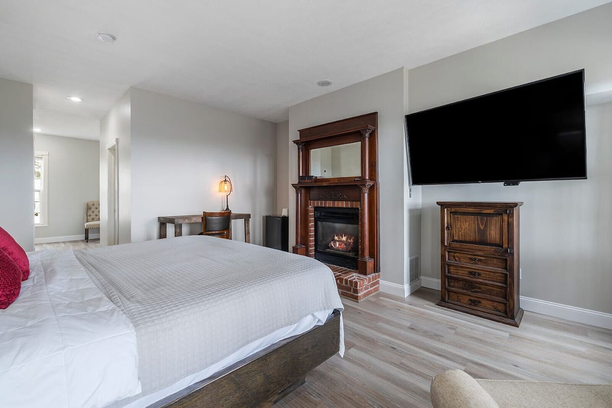 A bedroom with a large bed, a fireplace, a TV mounted on the wall, and a wooden cabinet.