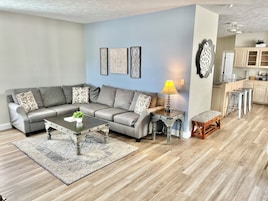 A living room with a grey sectional sofa, a coffee table, a patterned rug, and a small side table with a lamp.