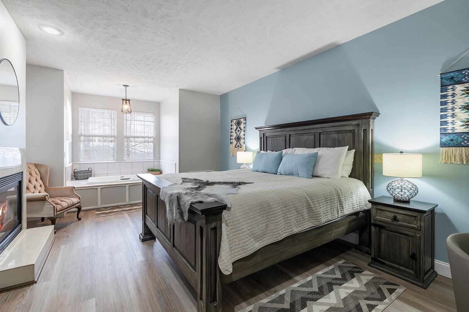 A bedroom with a large bed, a nightstand, a chair, a fireplace, and a window with blinds.