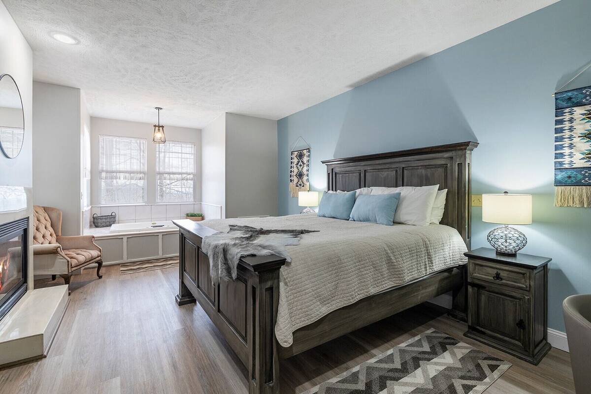 A bedroom with a large bed, a nightstand, a chair, a fireplace, and a window with blinds.