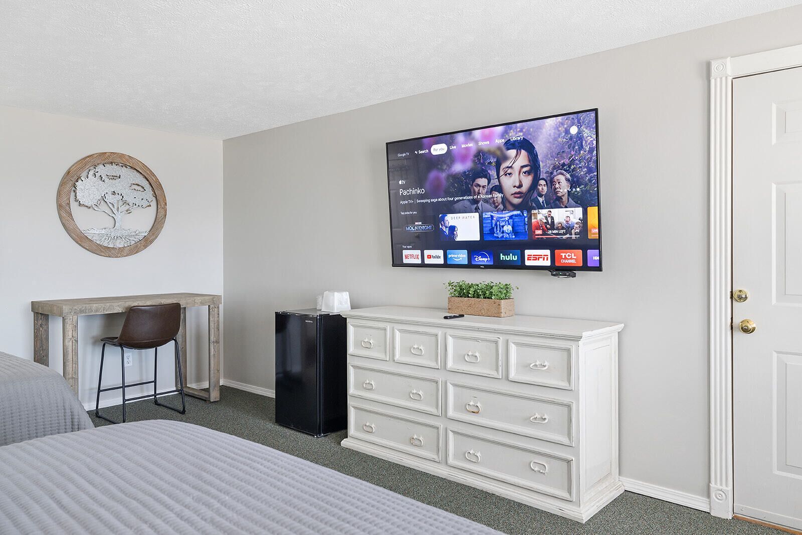A hotel room with a bed, a desk with a chair, a television mounted on the wall, a white dresser, and a door.