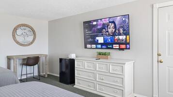 A hotel room with a bed, a desk with a chair, a television mounted on the wall, a white dresser, and a door.