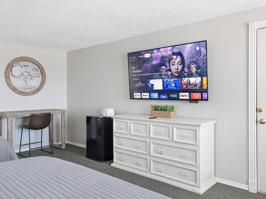 A hotel room with a bed, a desk with a chair, a television mounted on the wall, a white dresser, and a door.
