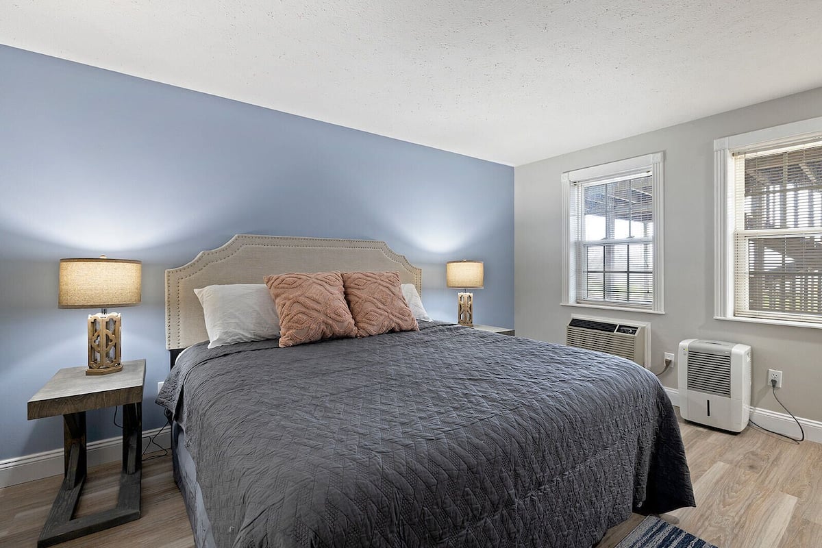 A bedroom with a bed, two bedside tables with lamps, a window with blinds, and an air conditioning unit.