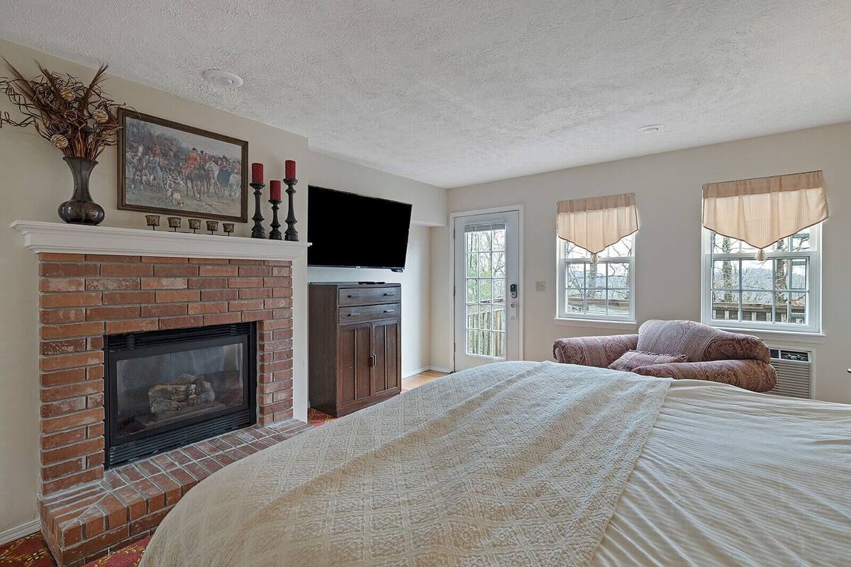 A bedroom with a brick fireplace, a large bed, a television, and a window with curtains.