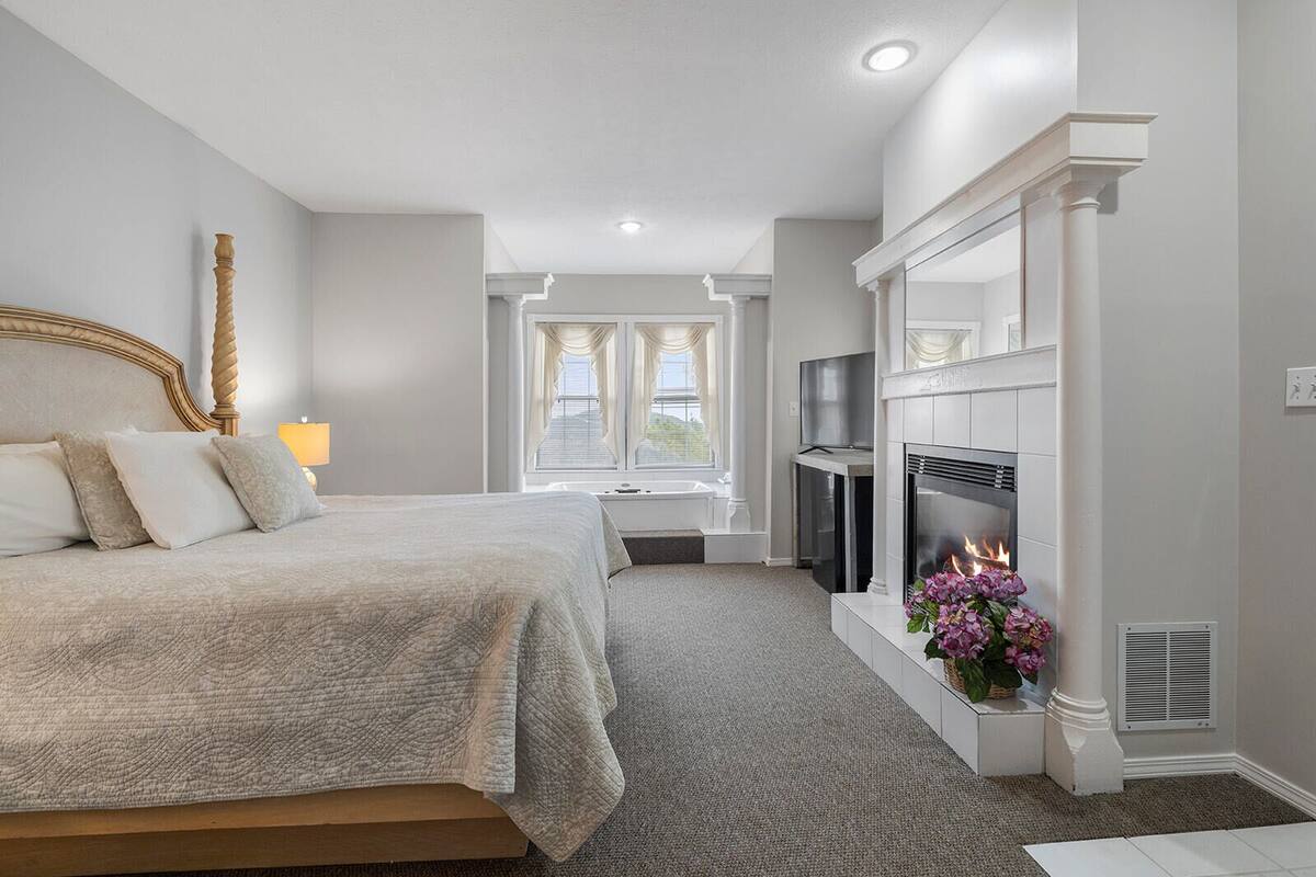 A bedroom with a large bed, a fireplace, a TV, and a window with curtains.