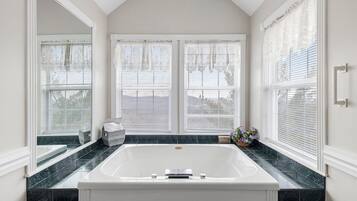 A bathroom with a large bathtub, two windows with white blinds, and a marble countertop.