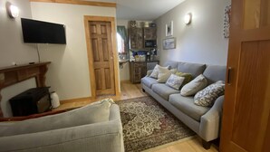 A living room with a grey sofa, a wooden door, a flat-screen TV mounted on the wall, and a kitchen area visible through an open door. - Adorable Tiny House in Picturesque Seaside Village (Warren)