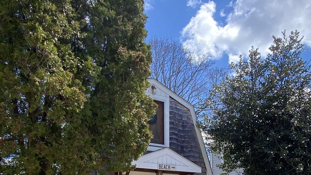 A two-story house with a wooden front door and a sign indicating a beach direction. - Adorable Tiny House in Picturesque Seaside Village (Warren)