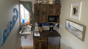 A compact kitchen with a sink, a microwave, and a coffee maker. There is a window with a curtain, a framed picture on the wall, and a small cabinet above the sink. - Adorable Tiny House in Picturesque Seaside Village (Warren)