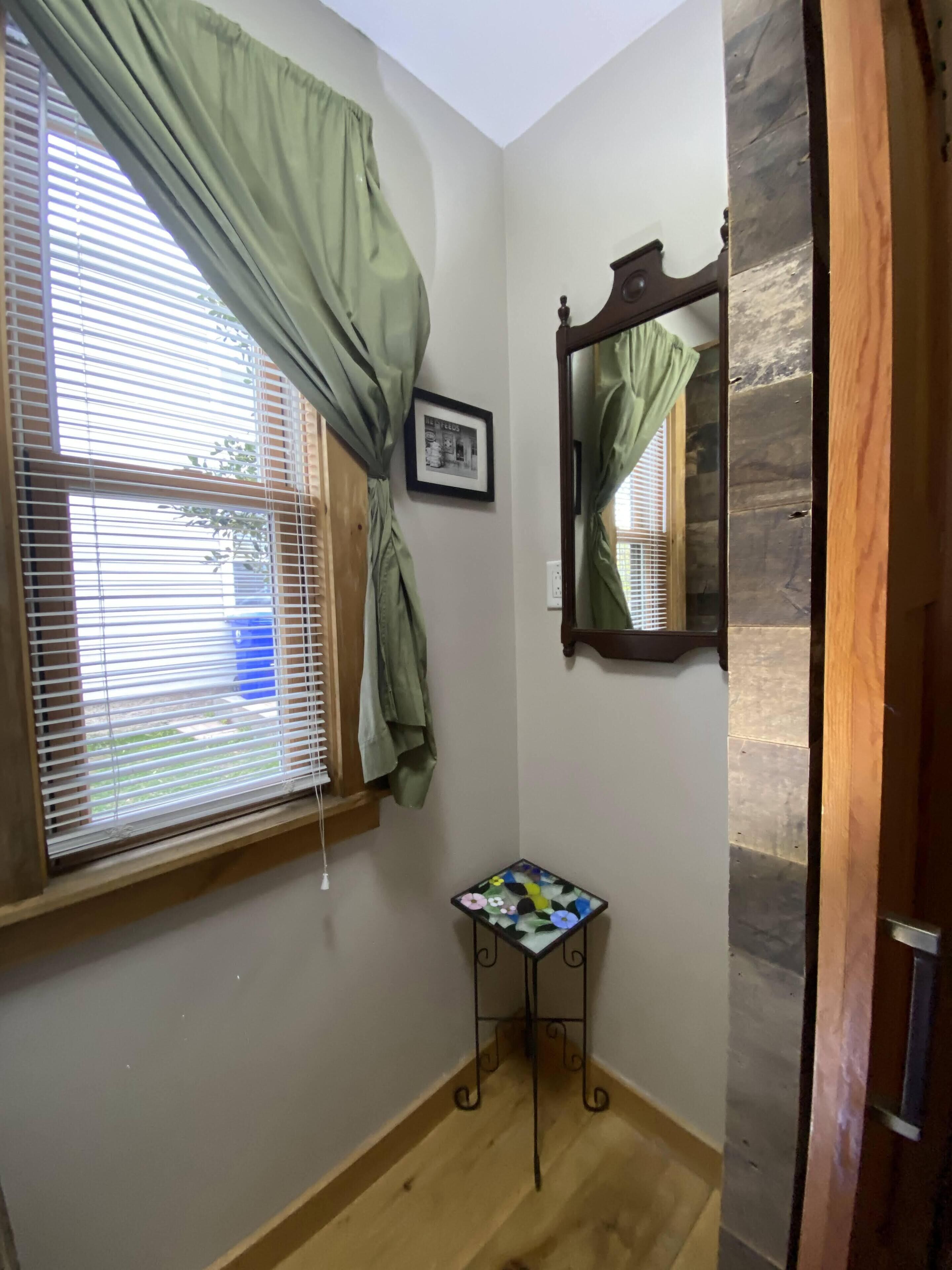 A small room with a window covered by green curtains, a framed picture on the wall, and a small table with a floral tablecloth.