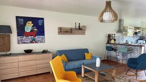 Ein Wohnzimmer mit einer Couch, Sesseln, einem Couchtisch, einem Gemälde, einer Küchenzeile und einer Pendelleuchte.