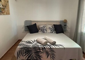 A neatly made bed with a white bedspread and two rolled towels, a headboard with horizontal slats, and a tropical leaf-patterned pillow. - Apartment (Le Gosier)