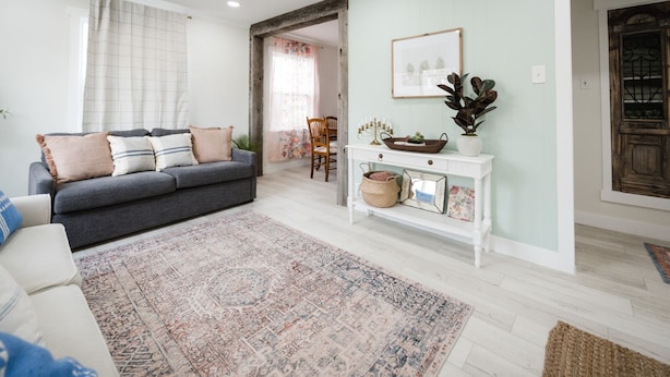 Un salon avec un canapé gris, un tapis à motifs, une console blanche et une porte ornée d’un panneau de verre décoratif.