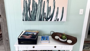 Une pièce avec un mur bleu clair, une console blanche, une œuvre d’art représentant un cactus encadré, un lecteur DVD, un livre et un panier décoratif contenant une plante.
