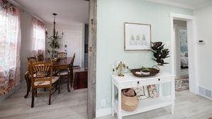 Un coin repas avec une table et des chaises en bois, une console blanche ornée d’un chandelier et d’un tableau encadré, et une porte-fenêtre donnant sur une autre pièce.
